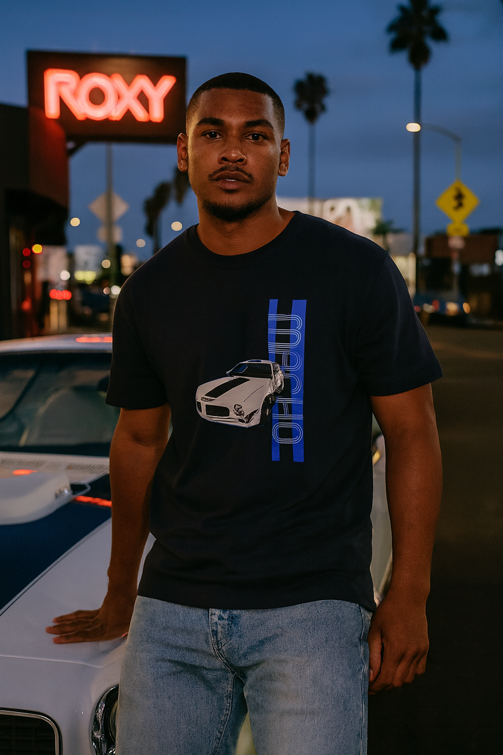 Man wearing a black bonspeed TransAm t-shirt with a car graphic and text, standing next to a white car at night.