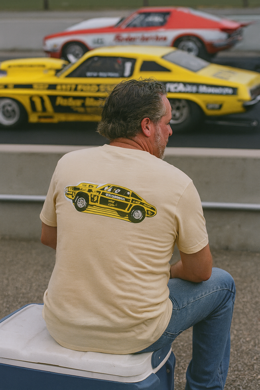 Man sitting on a stool wearing a bonspeed t-shirt with a race car design, with race cars in the background.