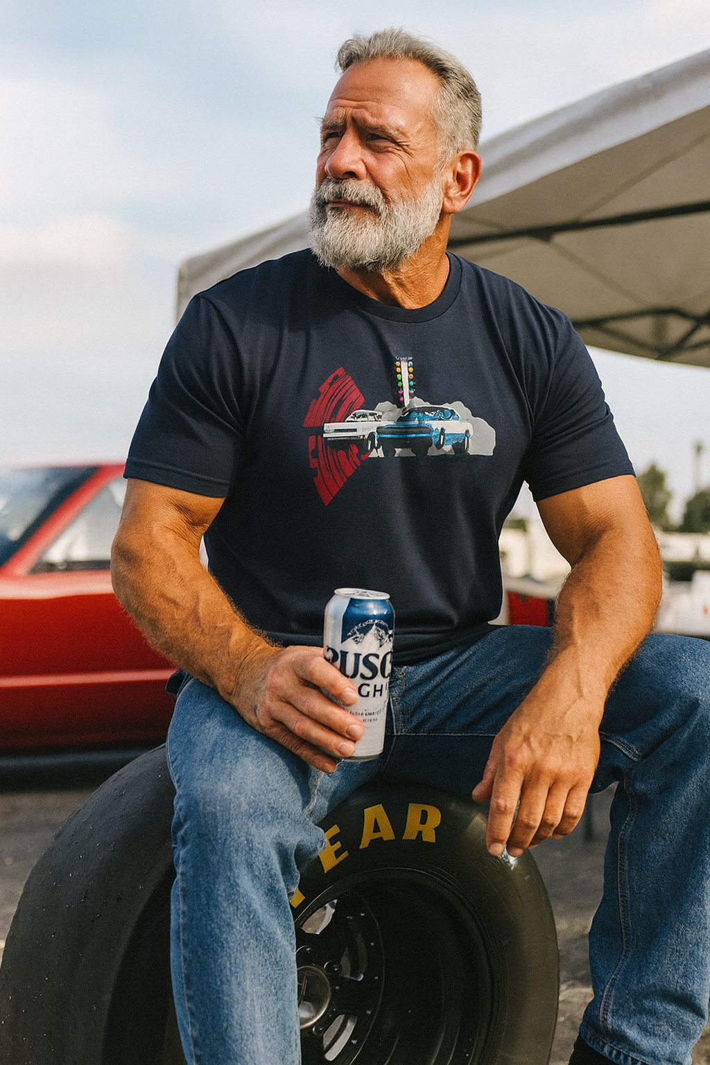 Man holding a beer can sitting on a tire with a truck in the background wearing a Pro stck Drag Racing bonspeed t-shirt 