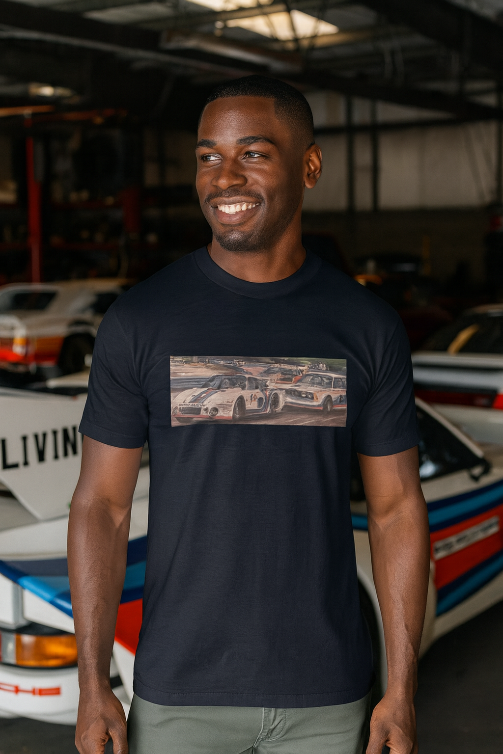 Man wearing a black t-shirt with a graphic Porsche BMW racing design, standing in front of vintage cars.