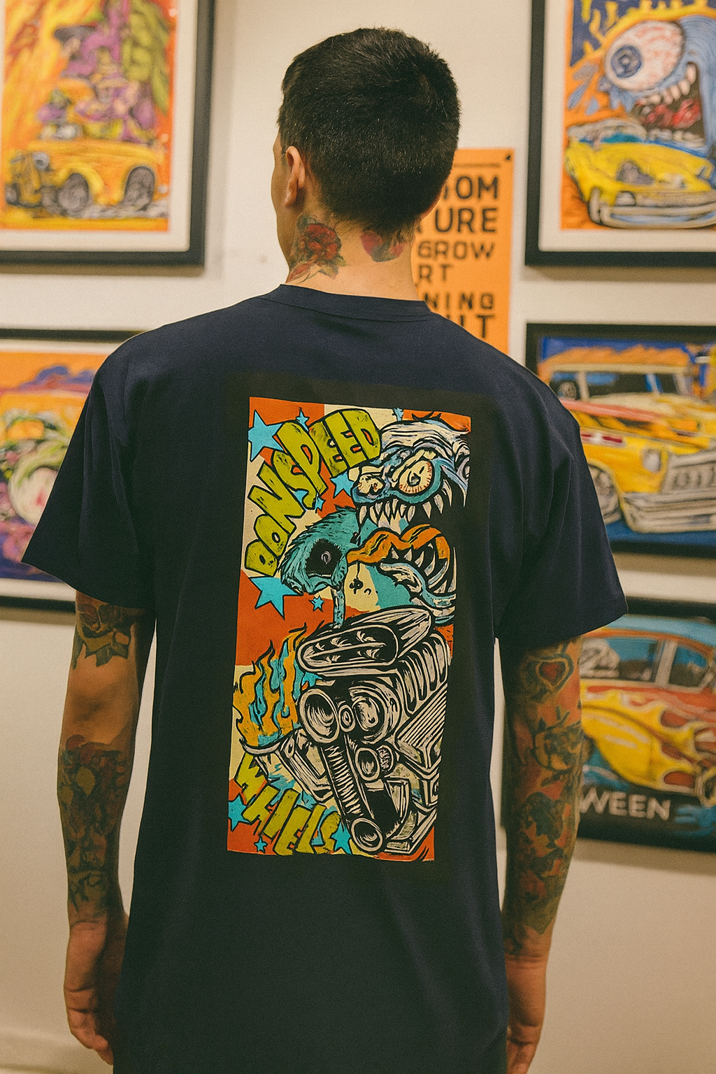 Person wearing a black bonspeed t-shirt with a colorful Rat Fink Style graphic design, standing in front of framed artworks.