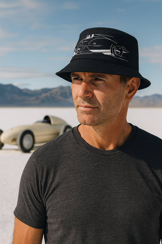 Man wearing a black bucket hat with a Bonneville car design, standing in a desert with a car in the background.