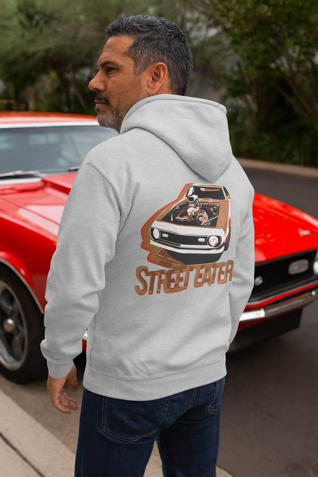 Man wearing a bonspeed hoodie with a car graphic and 'Street Eater' text, standing in front of a red car.